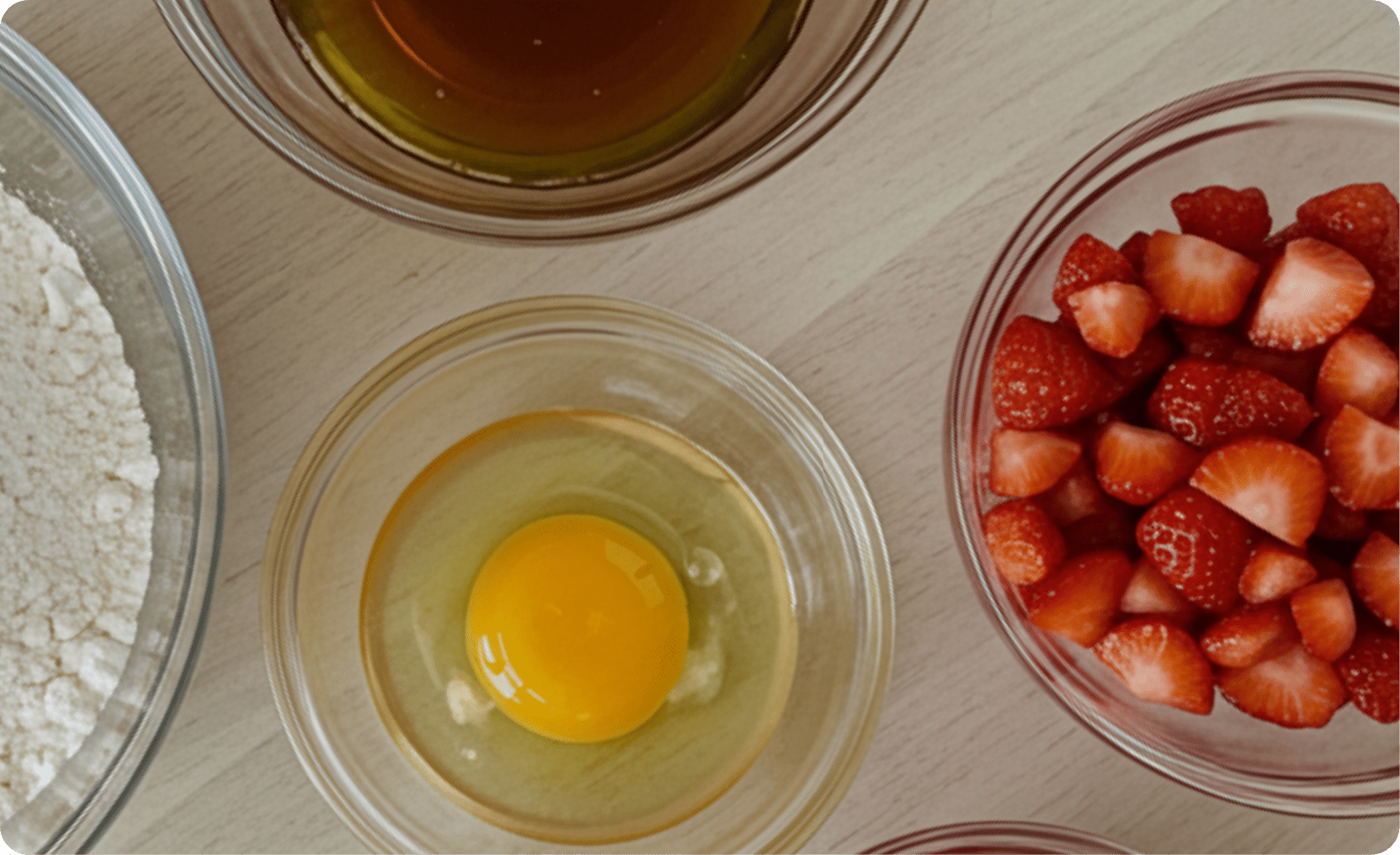 op-down view of baking ingredients including flour, raw egg, honey or syrup, and chopped strawberries in glass bowls.